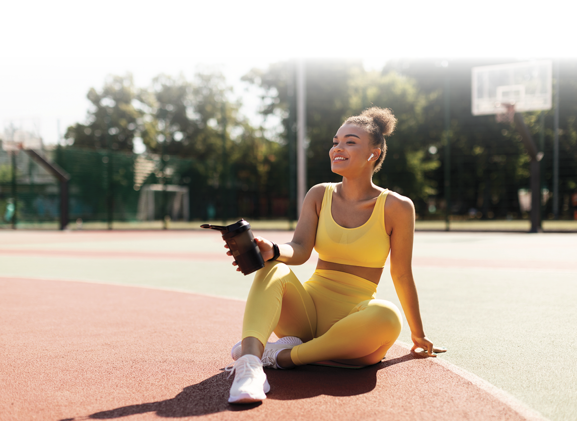 Energy, Vitality, Wellness Concept. Smiling African American woman sitting on basketball field ground, holding shaker, drinking water, taking break after outdoors exercise, wearing wireless earbuds